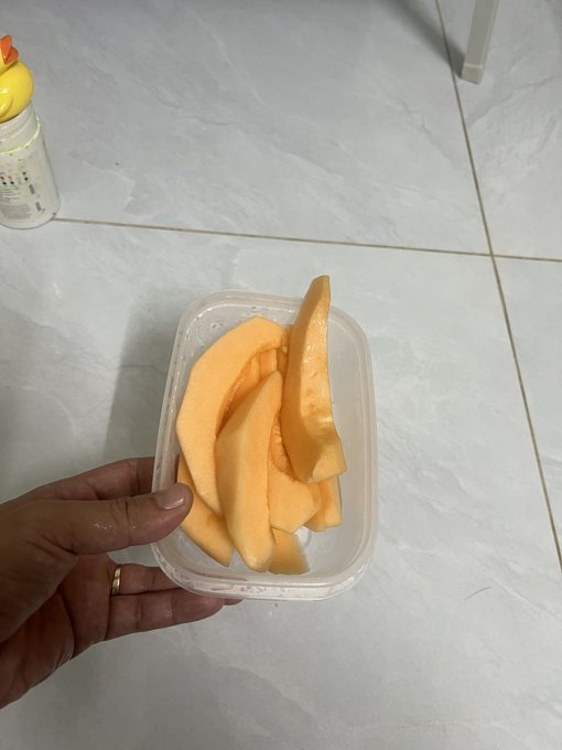 A hand holding a clear plastic container filled with sliced cantaloupe melon, photographed above a light-colored tiled floor with a baby bottle visible in the corner.