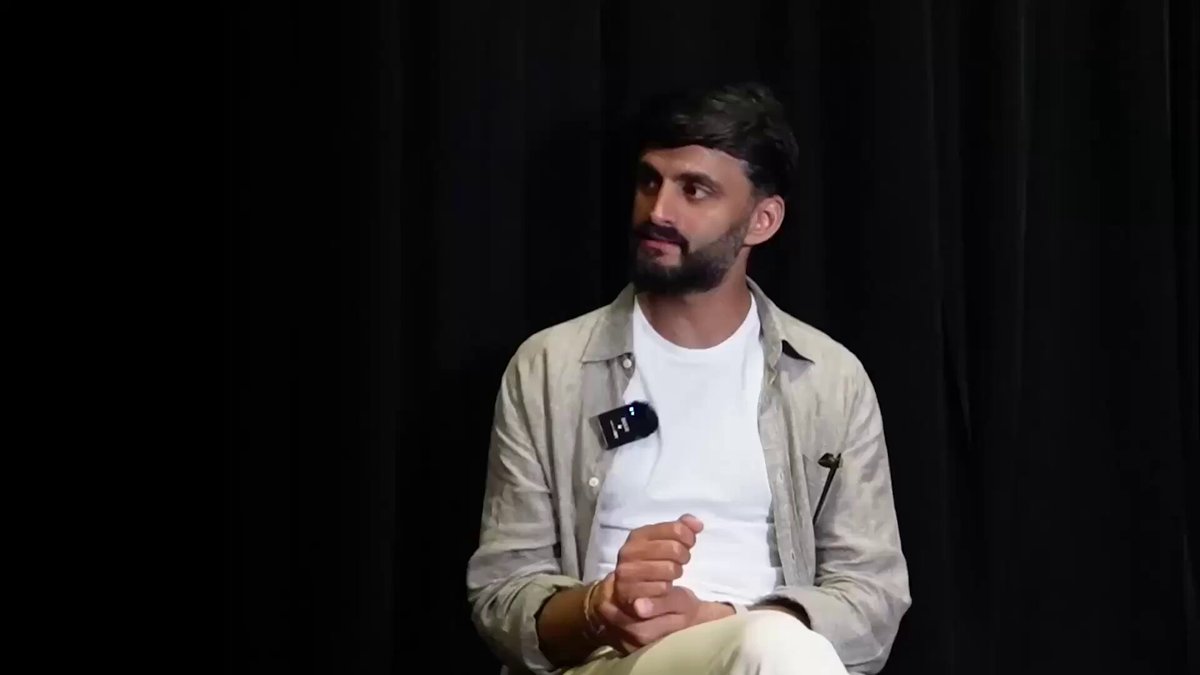 The image shows a man sitting down with his hands together while wearing a white shirt. The background of the image is black, which contrasts with the man's attire. There are no other objects or people visible in the picture.