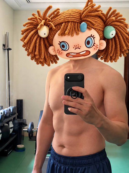 A shirtless man takes a mirror selfie in a gym, with a cartoonish doll filter featuring orange yarn hair placed over his face.