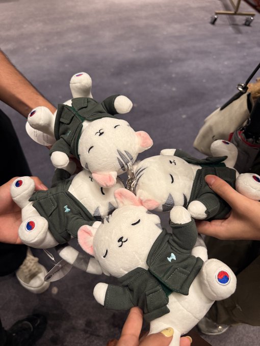 A group of hands holding up multiple plush toys featuring white cat-like characters wearing military-style green uniforms with South Korean flag patches.