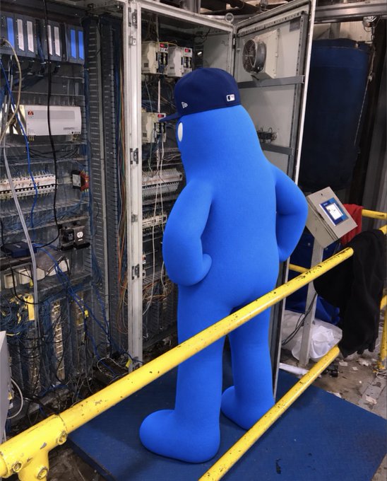 A blue mascot wearing a dark baseball cap stands confused amid open electrical panels and tangled wiring inside an industrial server room, humorously illustrating a chaotic first day on the job. The scene pairs with the post joking about starting work at @SlushWallet, capturing the mascot's cluelessness while inspecting the complex wiring setup.