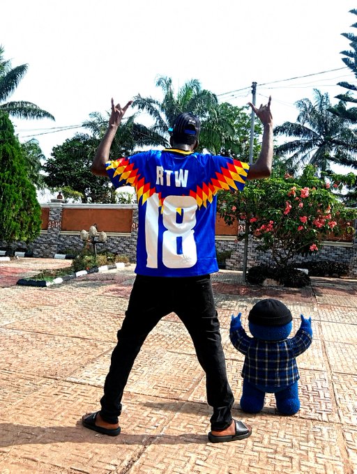 A person wearing a blue 'RTW 18' jersey stands with their back to the camera, raising both hands with two fingers extended, next to a small Smurf plush toy outdoors.