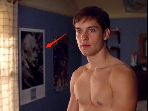 A young shirtless man with short brown hair standing in a bedroom, with a red arrow pointing to a black and white poster on the wall behind him that depicts an older man's face.