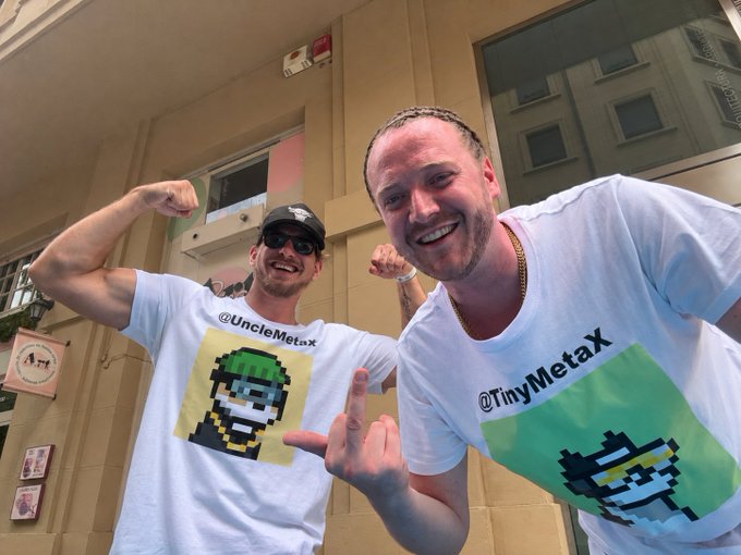 Two smiling men pose in front of a building, wearing white t-shirts featuring pixel art characters and the handles '@UncleMetax' and '@TinyMetax'.