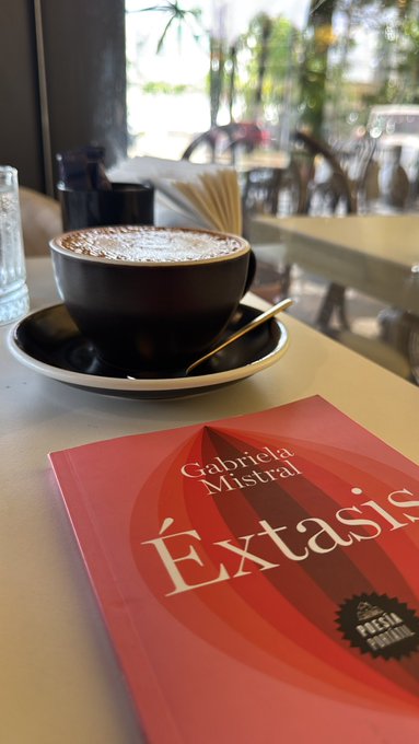 A black coffee cup on a saucer sitting on a table next to a red book titled 'Éxtasis' by Gabriela Mistral, with a 'Premio Pulitzer de Poesía' sticker.