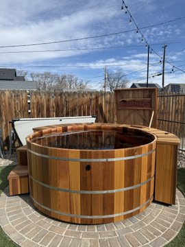 A wooden cedar hot tub sits in an outdoor backyard setting with a privacy fence and stone pavers, paired with a partial view of a white cover. The caption "Done - bitcoin though." suggests the hot tub installation is complete, though the Bitcoin reference humorously implies the cost of the project drained the owner's cryptocurrency funds.