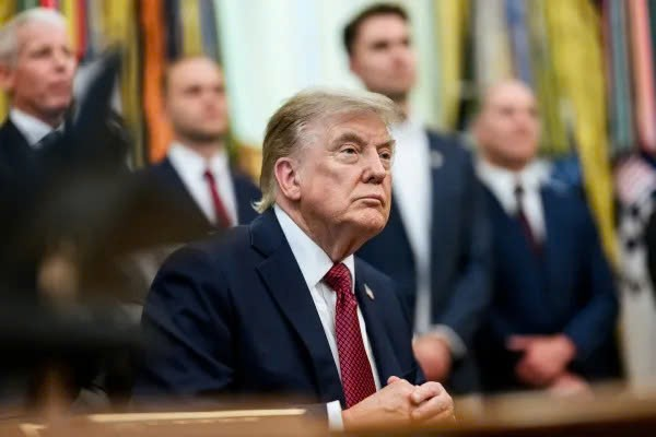 Donald Trump is seated at a table in a formal setting, wearing a dark suit and red tie with a serious expression, while four men in suits stand in the blurred background.