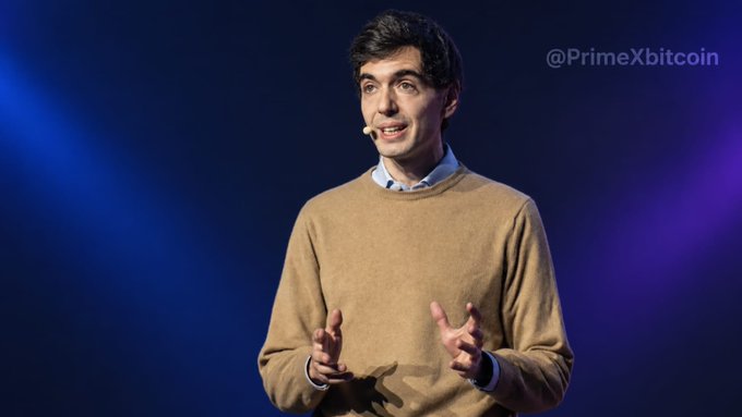 The image shows a man with dark hair, wearing a tan sweater over a collared shirt, speaking on stage with a microphone headset against a blue-lit backdrop, illustrating a tech/corporate presentation context relevant to Stripe's expansion of stablecoin payment services to over 100 countries. The watermark "@PrimeXbitcoin" appears in the upper right corner.