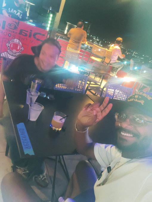 Two people pose together at an outdoor nighttime venue, with drinks on a table and a 'LIQUID DEATH' sticker visible on the table edge.