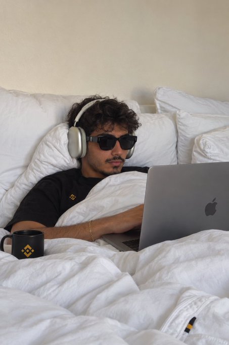 A man wearing sunglasses and headphones reclines in white bed linens while browsing on his Apple MacBook, accompanied by a black Binance-branded coffee mug—the Binance logo ties into the caption "One last look… and then sleep," suggesting a final crypto market check before bed.