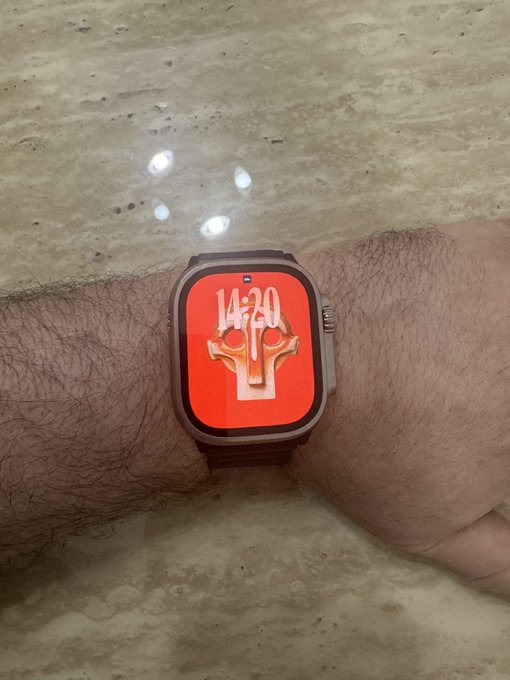 A close-up of an Apple Watch on a wrist displays a red watch face featuring a stylized gold cross design and the time '14:20', resting over a stone surface.