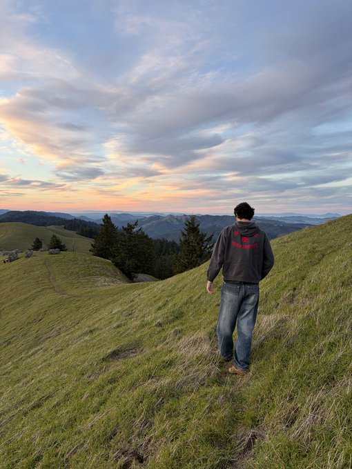 The image features a man walking up a grassy hill, with a beautiful sunset behind him. The sky has a mix of orange and blue hues, creating a stunning backdrop for the scene. The man appears to be enjoying his time outdoors as he takes in the view from the top of the hill.