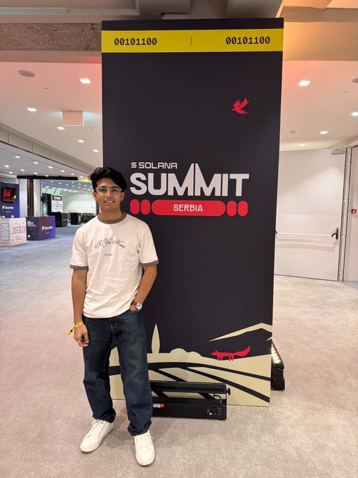 A young man in glasses and a white t-shirt poses in front of a tall black banner reading "SOLANA SUMMIT SERBIA" with the code "00101100" displayed at the top, standing inside the convention venue where the event took place.
