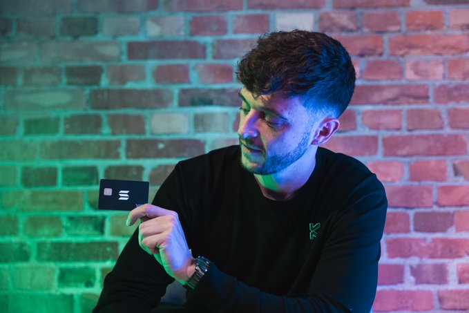 A young man with short brown hair and a beard, wearing a black long-sleeve shirt, looks down thoughtfully while holding a black card featuring a contactless payment symbol, illuminated by blue and green lighting against a brick wall.