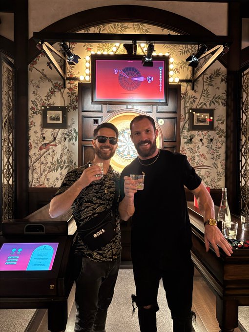 Two men pose with drinks in an ornately decorated room featuring a dartboard and screens, promoting a 9:00pm EST live stream hosted by @doodlegenics and @kokid951 on @bcgame, focused on memecoins, parlays, and gaming.