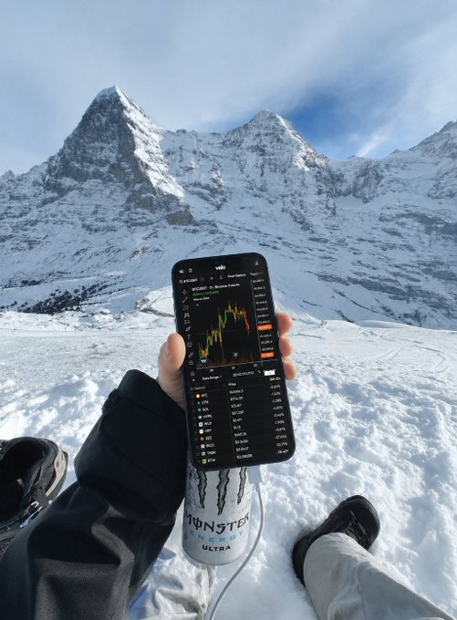 The image features a person wearing a black jacket sitting on top of some snow, holding up their phone with a graph displayed on its screen. The graph appears to be a chart depicting stock prices or other financial data related to energy companies. This scene suggests that the individual is interested in monitoring and analyzing such information from a remote location while being outdoors, possibly during wintertime.