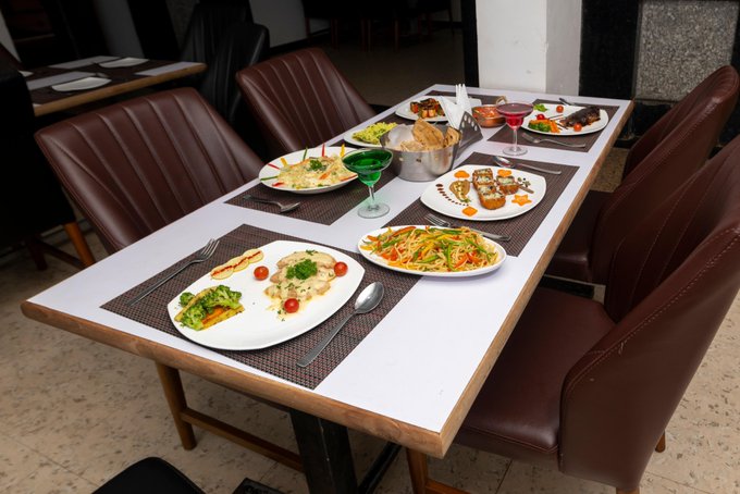 A restaurant table is set with multiple plates of food, including salads, rice, and meat dishes, alongside wine glasses and brown leather chairs.