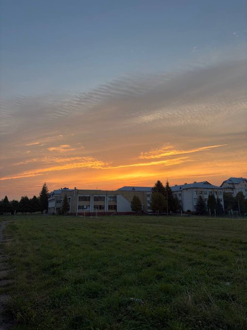 A serene sunset paints orange and pink hues across the sky above a long, low-rise apartment or school building surrounded by an expansive grassy field. The peaceful, scenic view reflects the post's introspective tone about finding direction and carving out a unique path for the account.