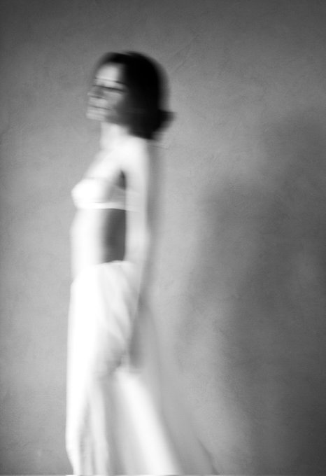 A black-and-white artistic photograph of a woman in a long white dress standing in profile, with motion blur creating a ghostly, dreamlike effect against a textured gray background.