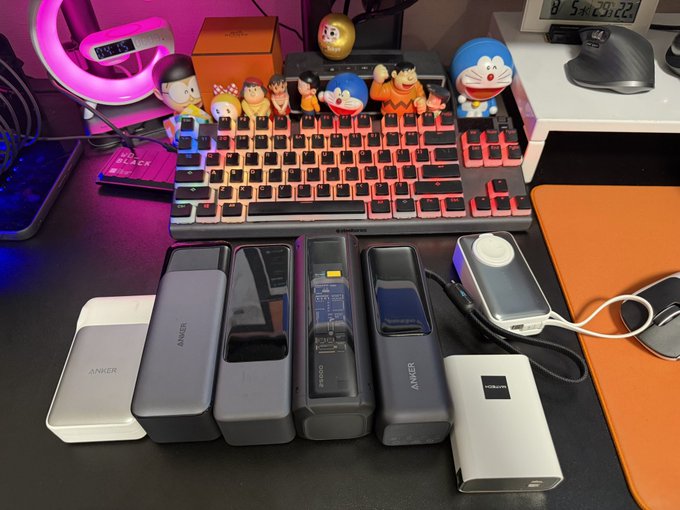 A desk features a colorful backlit keyboard lined with cartoon character figurines, alongside six Anker-branded power banks and one CUKTECH unit arranged in front. However, looking carefully, the sixth Anker-branded device on the left appears to actually be a different brand, contradicting the visual expectation.