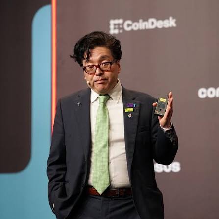 A man wearing glasses, a dark suit, and a green tie gestures while speaking on stage at a CoinDesk Consensus event, with the "CoinDesk" logo visible in the background, contextualizing the announcement of BitMine's $19 million Ethereum purchase of 10,399 ETH amid Wall Street's growing ETH accumulation trend.