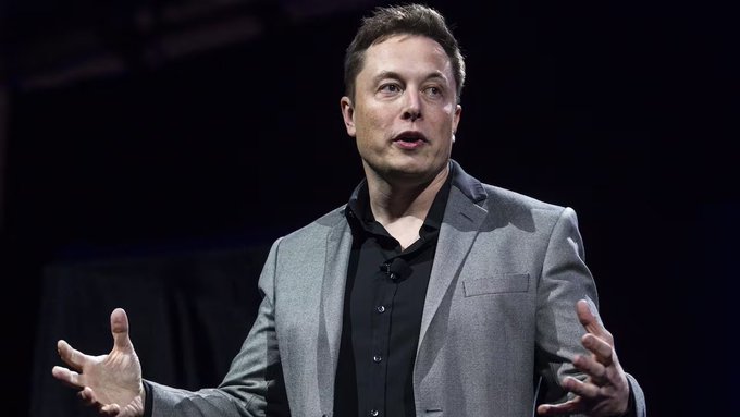 Elon Musk speaking on stage, wearing a gray blazer over a black shirt, gesturing with both hands open.