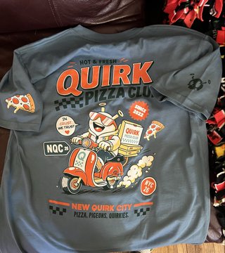 A grey Quirk Pizza Club t-shirt featuring a cartoon cat riding a scooter with the text "New Quirk City" and "Hot & Fresh."