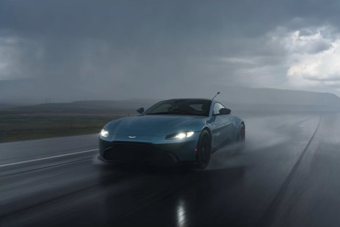A blue Aston Martin sports car driving on a wet road in rainy, overcast conditions with headlights illuminated.