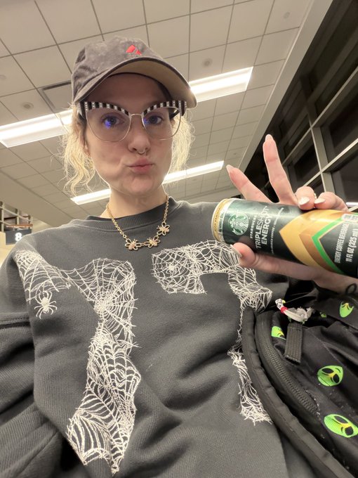 A person wearing a brown cap, round glasses, and a gray sweatshirt with white spiderweb designs, holding up a peace sign and a can of 'Triple Shot Energy' drink.