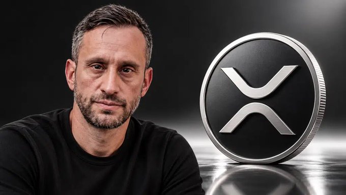A man with short dark hair and a beard stands next to a large physical representation of the XRP cryptocurrency coin.