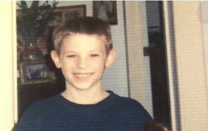 A young boy with short brown hair smiles at the camera, wearing a dark blue shirt in what appears to be a home setting.