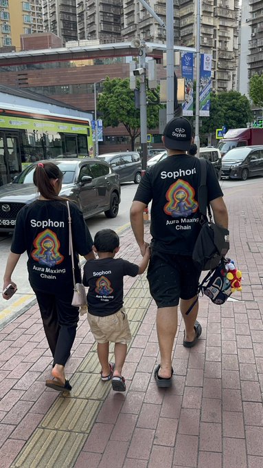 The image shows a man, woman, and child walking down a city street together. The man is wearing a black t-shirt with the name "Sophon" printed on it, while the woman has a backpack on her back. A young boy is also present in the scene, holding onto his mother's hand as they walk along the sidewalk.

The image appears to be related to a post or social media account that features a countdown for the release of a new movie called "Touching Grass on a beautiful Sunday!". The caption suggests that this event will take place after the release of the movie, and it seems like an exciting time for fans of the film.