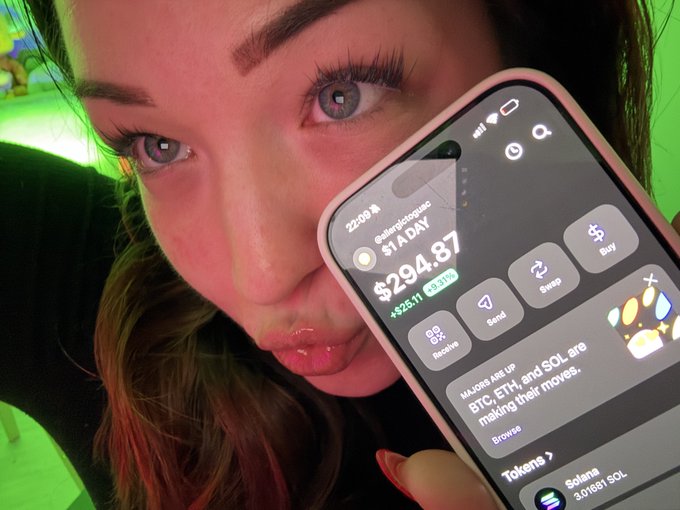 A woman holds up her phone displaying a crypto portfolio showing a $294.87 balance with a +$25.11 gain (+9.31%) on Solana (SOL), with her current holdings of 3.01691 SOL visible at the bottom. The image captures her excitement as her day 275 daily $1 Solana buying strategy finally turns profitable, aligning with the post's celebration of reaching "IN PROFITTTT" territory.