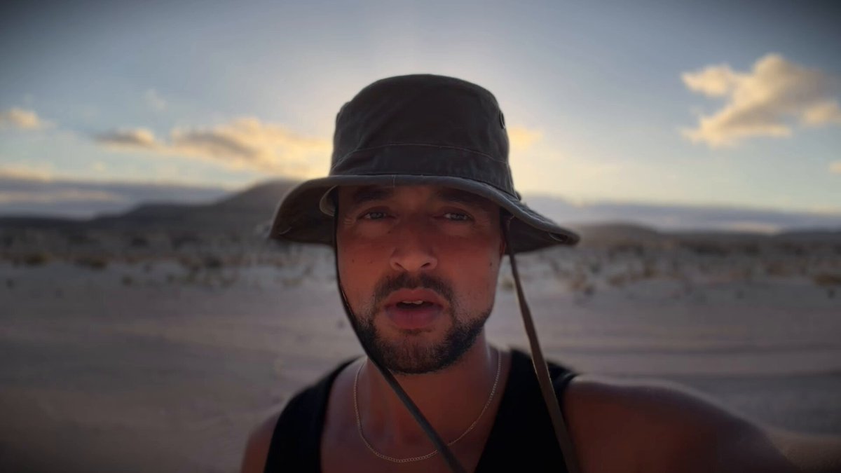 The image features a man wearing a hat, standing in an open area with mountains in the background. The man appears to be looking directly at the camera while holding something in his hand. There are no other people or objects visible in the scene.