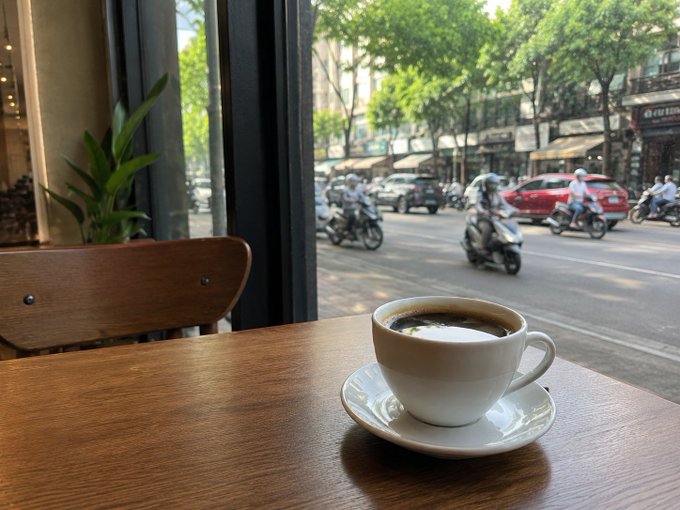 A steaming cup of black coffee sits on a wooden table beside a window, overlooking a bustling street filled with motorbikes and cars as the city wakes up — capturing the quiet morning ritual that contrasts with the busy traffic outside.