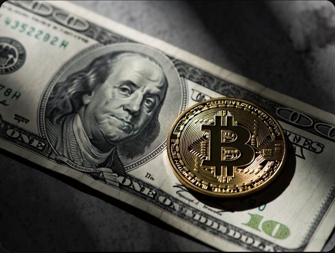 A gold Bitcoin coin resting on top of a US one hundred dollar bill featuring Benjamin Franklin's portrait.