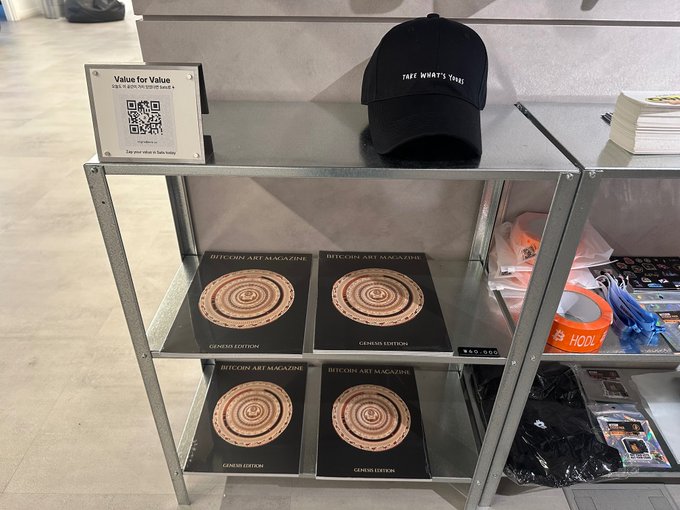 A retail display shelf holds copies of "BITCOIN ART MAGAZINE GENESIS EDITION," a black "FREE WHATS THINGS" cap, and merchandise accessories, with a "Value for Value" QR code sign on top.