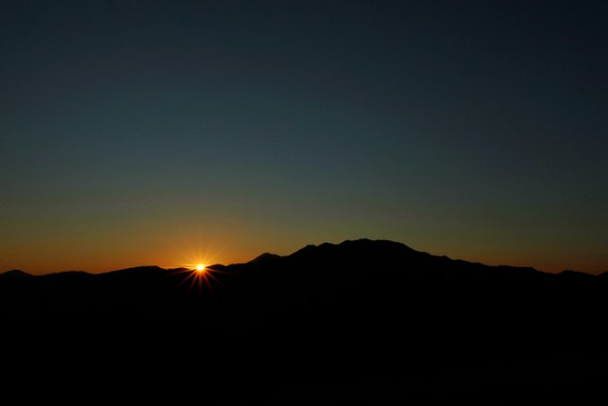 A scenic sunrise photograph showing the sun peeking over a dark mountain silhouette against a gradient sky transitioning from orange/yellow near the horizon to deep blue above.
