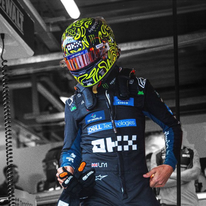 A photo of Formula 1 driver Lando Norris (identified by the 'LN4' British flag patch) in his McLaren racing suit and a brightly patterned yellow and green helmet, standing in a garage or pit area.