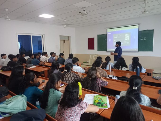 A classroom scene where students are seated at wooden desks facing a professor who is giving a presentation displayed on a projector screen.
