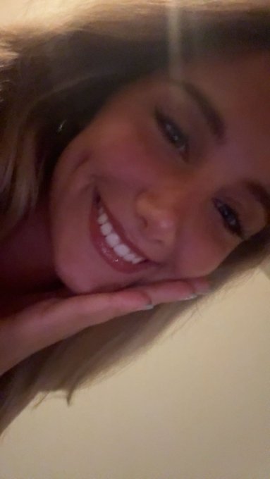 A close-up selfie of a smiling blonde woman with dark eyeliner and pink lipstick, resting her chin on her hand against a white pillow.