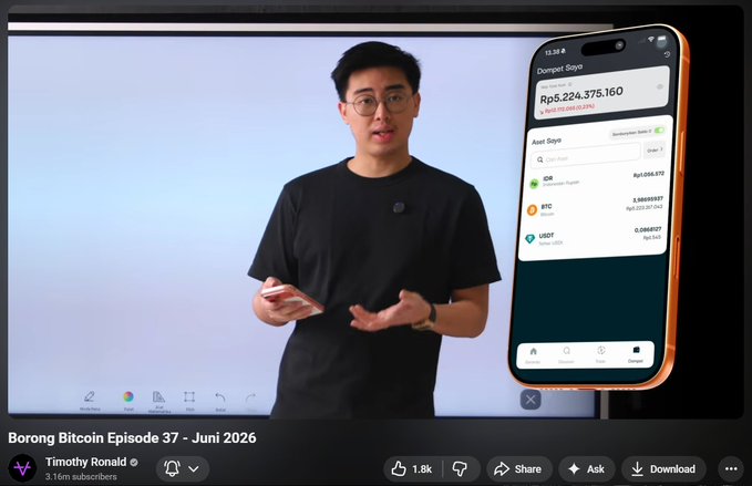 The image shows a man standing in front of a computer screen, holding up an iPhone with a video playing on the screen. The man appears to be giving a presentation or discussing something related to Bitcoin. There are several other elements in the scene that contribute to the overall atmosphere and context of the image.