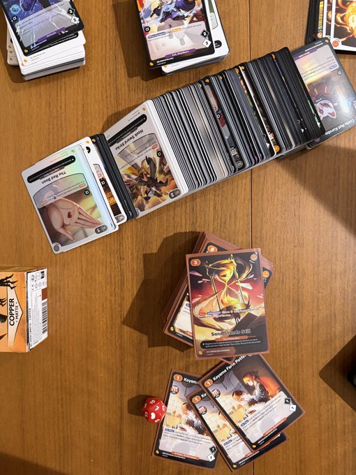 A collection of trading/collectible cards spread across a wooden table, with one card prominently showing 'Savage Flame Burst' artwork, alongside a red d20 die and a card box labeled 'COPPER'.