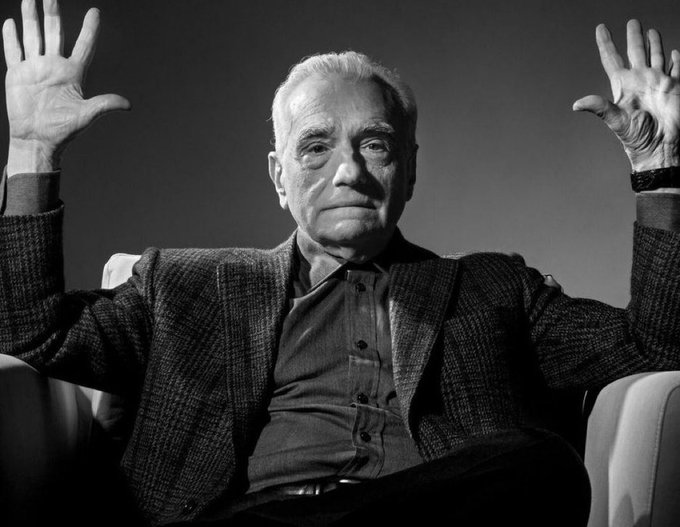 A black-and-white portrait of an elderly man in a tweed jacket sitting in a chair with both hands raised.