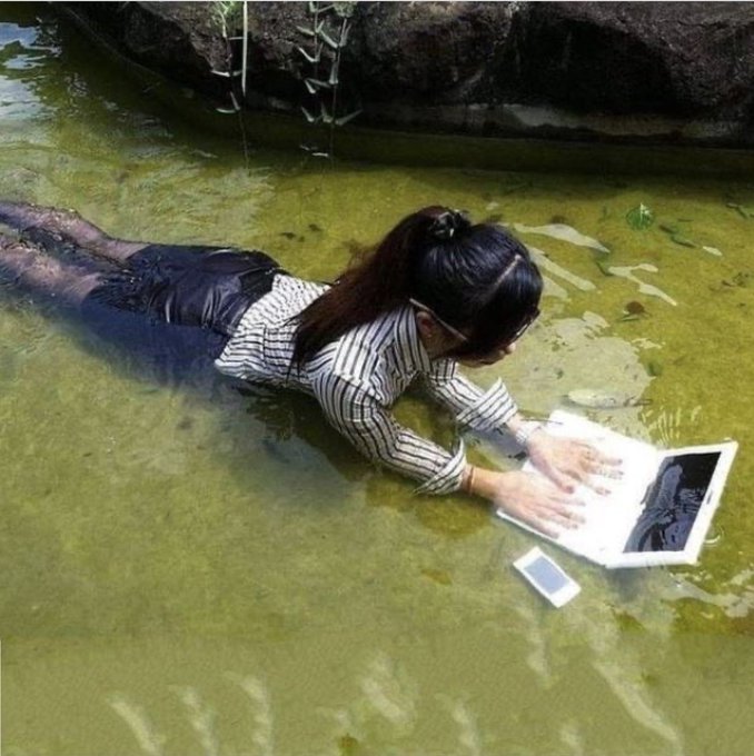 A woman in a striped shirt and dark skirt lies on her stomach in shallow, murky green water, holding an open book and a smartphone in front of her, appearing to read while submerged.