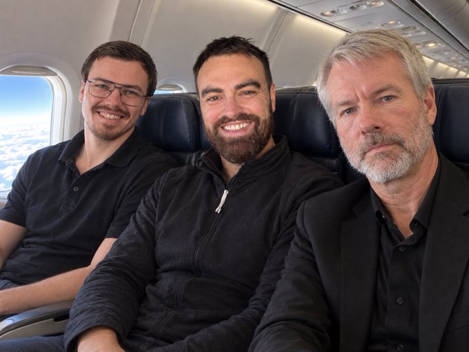 Three men sit side-by-side on an airplane, with a younger man on the left smiling, a bearded man in the middle grinning broadly, and an older man with gray hair on the right.