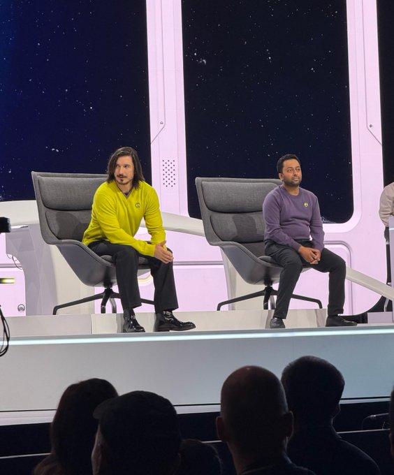 Two men sit on a futuristic stage with a starry space background backdrop, one wearing a bright yellow sweater and the other in a purple sweater.