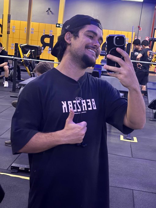 A selfie of a smiling young man in a gym wearing a black 'BERZERK' t-shirt and a backward cap, giving a thumbs up while holding a smartphone.