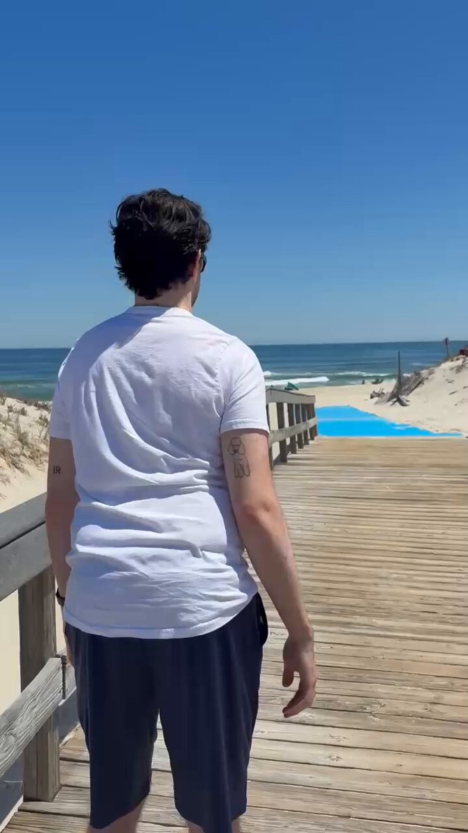 The image features a man standing on a wooden boardwalk, looking out at the ocean with his back turned towards the camera. The man appears to be wearing a white t-shirt and black shorts while enjoying the view of the beach. There is also a blue mat placed near him, possibly for additional comfort or safety as he walks along the boardwalk.