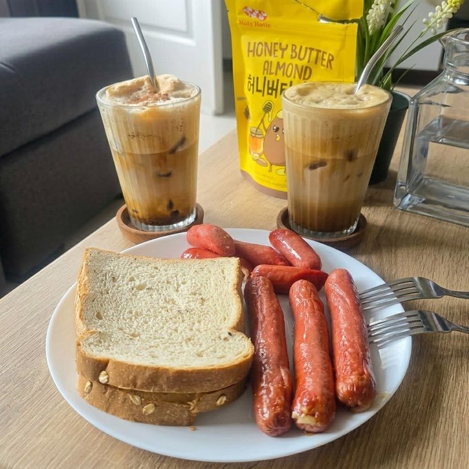 A breakfast spread on a wooden table featuring a plate of toasted bread and grilled sausages alongside two glasses of iced coffee and a yellow pouch labeled "HONEY BUTTER ALMOND."